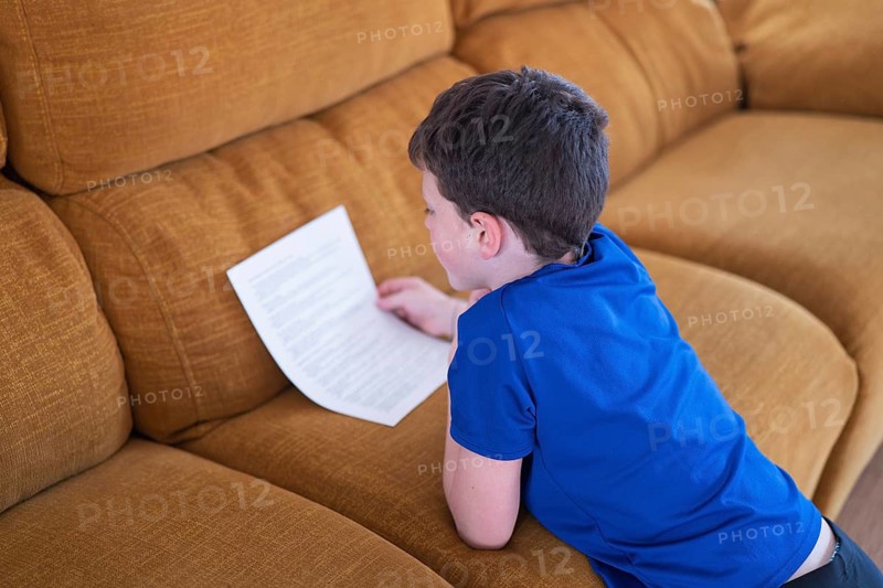 A boy in a blue shirt attentively reads a sheet of paper while sitting on a comfortable couch
