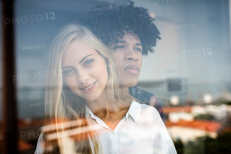 A reflective image of a diverse couple gazing through a window. Natural light softly illuminates their faces, creating a serene and thoughtful atmosphere