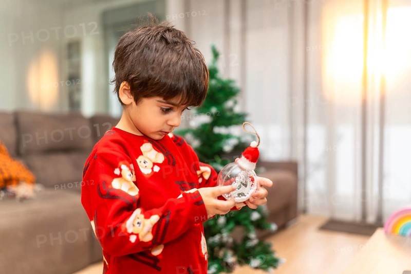 Young boy in cozy pajamas studies a clear christmas ornament topped with a tiny santa hat as he prepares to decorate the family tree, warm natural light filling the room