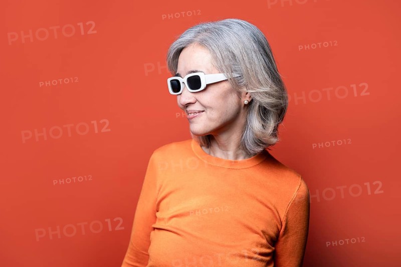 Asian Chinese senior woman with grey hair wearing trendy white sunglasses and an orange sweater. She stands against an orange background, embodying confidence and style