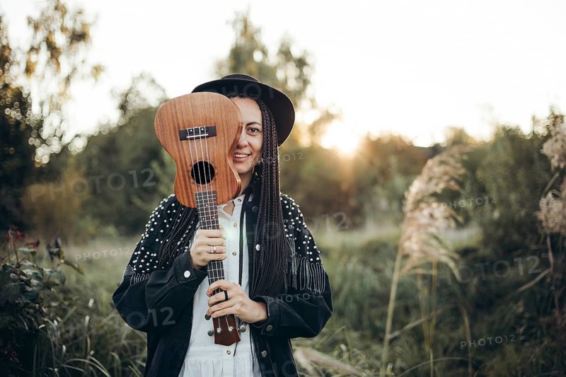 A woman stands outdoors holding a ukulele, enjoying the peacefulness of the sunset. The warm glow creates a serene atmosphere, emphasizing the beauty of nature and music