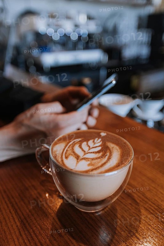 A man uses a smartphone at a cafe, focusing on a beautifully crafted latte with intricate art. The warm ambiance enhances the cozy, relaxed atmosphere