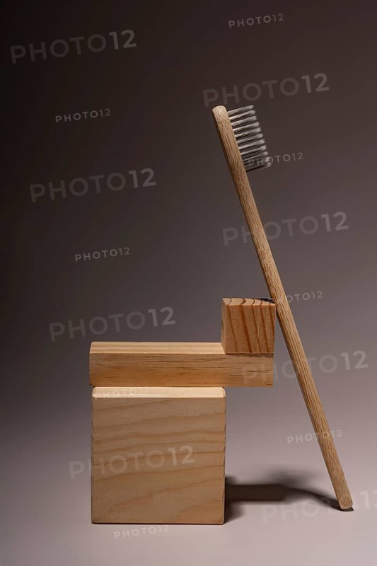 A bamboo toothbrush stands against a wooden block backdrop on a soft brown surface. The natural materials and muted colors create an eco-friendly aesthetic, ideal for sustainable living