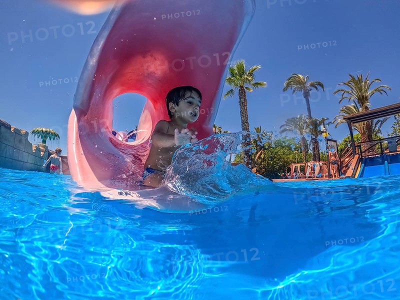 Happy child exiting a pink waterslide and splashing into a blue swimming pool at a water park, enjoying summer vacation