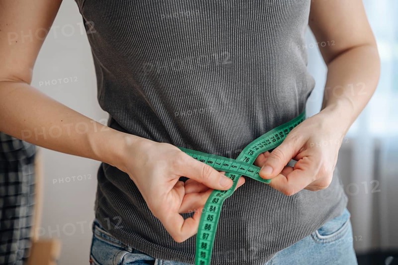 A woman measures her waist with a green tape measure, symbolizing her weight loss journey. She wears a casual outfit, embodying commitment to a healthy lifestyle