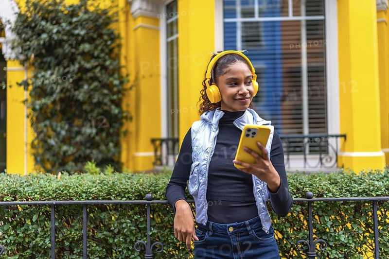Portrait of a cool african american woman listening to music using phone leaning on a rail next to yellow facade house