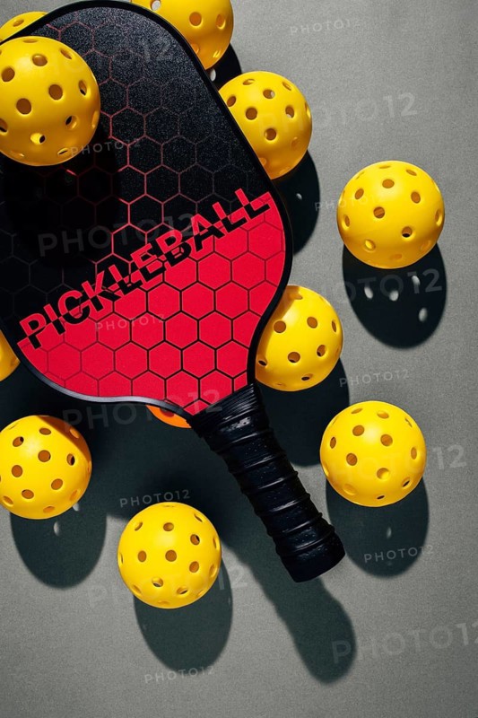 Top view of a pickleball paddle with a red and black design surrounded by vibrant yellow balls. Set against a gray background, this image captures the essence of modern sports gear