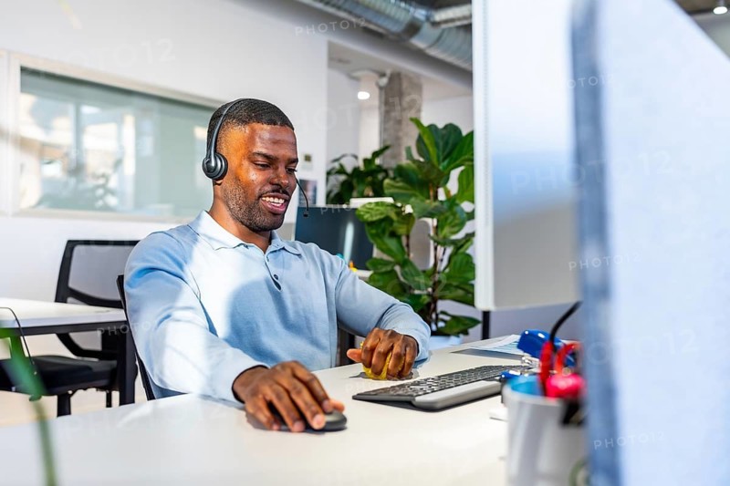 Young black man working at a computer while wearing a headset in a bright, modern coworking office, focused on delivering customer support