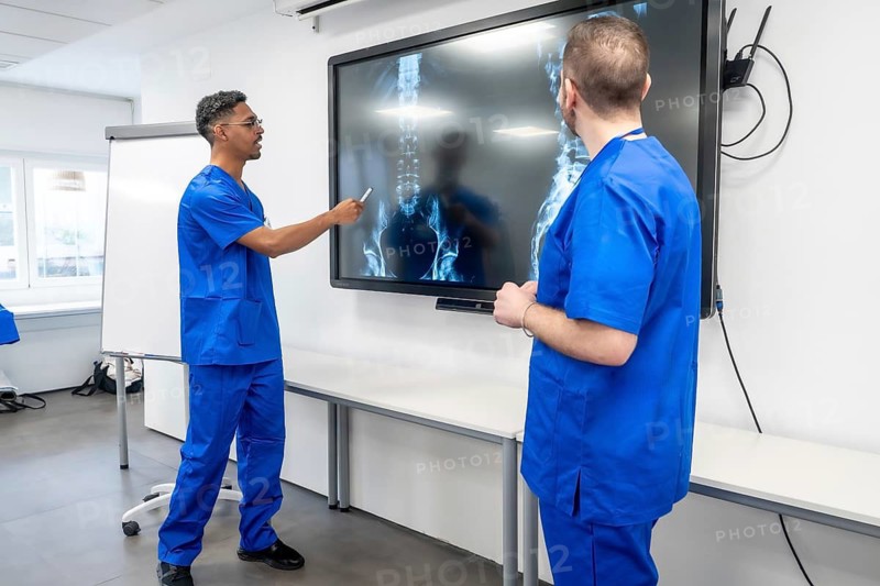 Two medical professionals wearing blue scrubs discussing a patient's spinal x rays displayed on a large digital screen, focusing on diagnosis and research in a modern healthcare setting