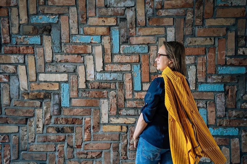 Side view of a 40-year-old woman in glasses against a colorful brick wall, Woman in casual attire stands by a patterned brick wall with bright colors