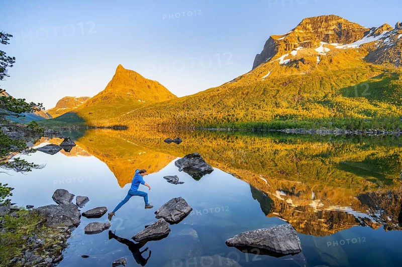 Young man jumping from stone to stone in water, picturesque mountain landscape in the Innerdalen high valley with Innerdalstårnet summit, mountain peaks reflected in Lake Innerdalsvatna, in the evening light at sunset, Trollheimen Mountain Area, Sunndal, Møre og Romsdal, Norway