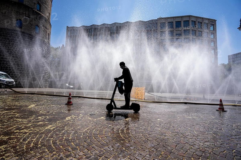 Cooling Cologne, campaign by the city of Cologne to cool down places exposed to heat, here Rudolfplatz in the city center, at just under 37°C during the day, a spray hose splashes fine water droplets that cool down the immediate area a few degrees, an action carried out when there is extreme heat stress, to adapt the urban climate to climate change, passers-by enjoy the cooling moment, plus tips on how to deal with extreme Hitze, North Rhine-Westphalia, Germany