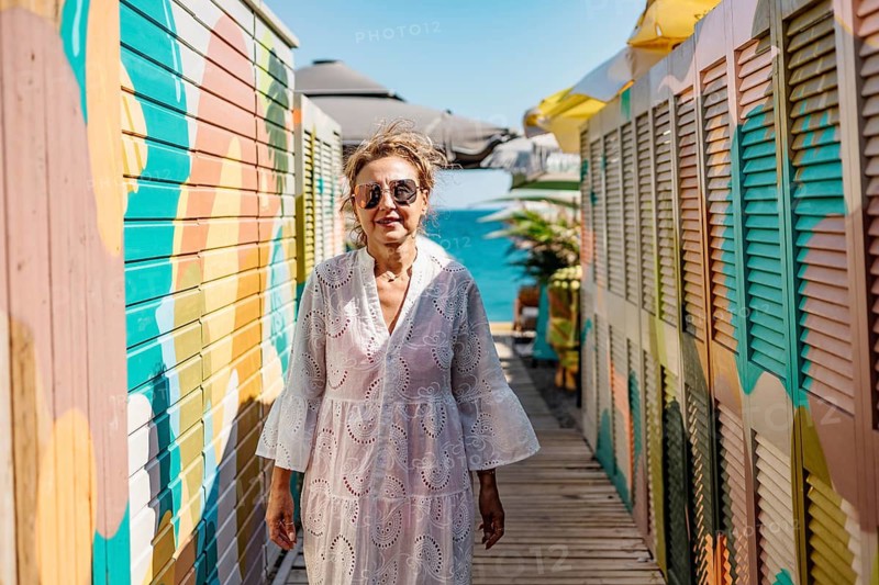 Senior Caucasian woman with glasses walks along a colorful boardwalk by the water. She wears a white dress and enjoys a sunny day