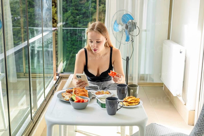 A young Caucasian woman with long brown hair sits at a table on a balcony. She is using a smartphone while enjoying a meal with various dishes and drinks