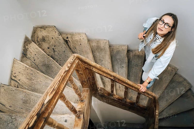 A woman looks up standing on an old wooden staircase, Woman with glasses on a wooden spiral staircase, looking up, creating an introspective atmosphere
