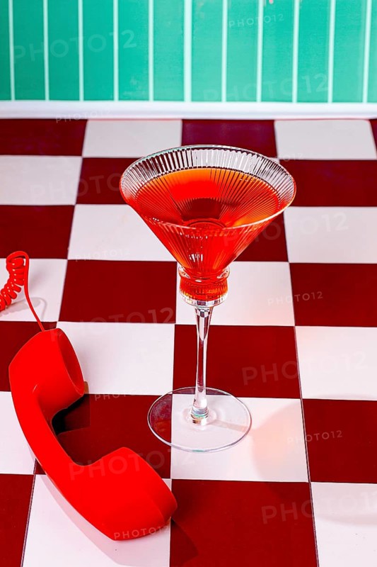 A glass filled with a red drink is placed on a table. A red phone sits beside the glass. The walls are covered with green tiles. The floor has a checkered pattern