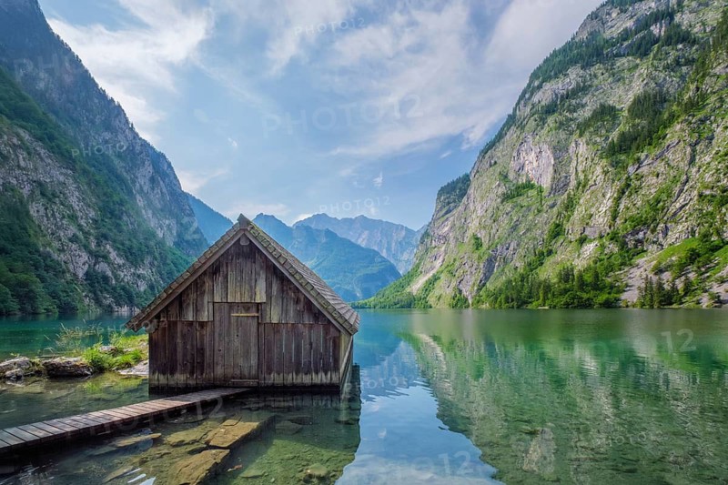 Wide shot of a wooden boat shed on the calm Obersee lake under a blue sky with soft clouds, Bavarian Alps, Germany