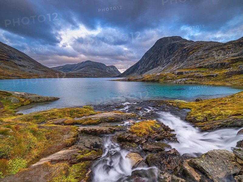 A mountain lake with a small waterfall surrounded by rocks and a dramatically cloudy sky, sunset Lake Djupvatnet, autumn, mountain, Geiranger, Møre og Romsdal, Vestland, Norway