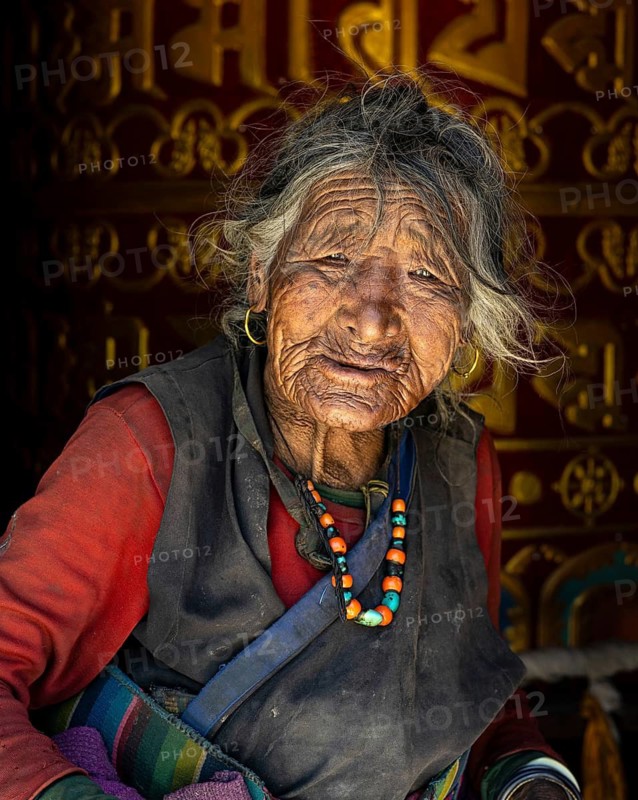 Very old woman with coral and turquoise necklace in traditional clothing in front of giant Buddhist prayer wheel, Choser, Mustang, Upper Mustang, former kingdom, Himalayas, Nepal