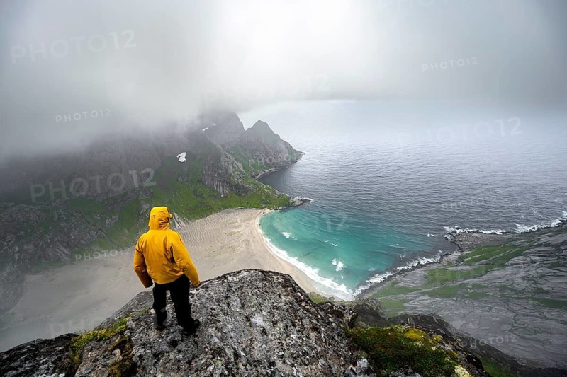 Hiker on Helvetestinden summit, view of Bunesstranden sandy beach and blue sea, dramatic cloudy sky, Lofoten, Norway