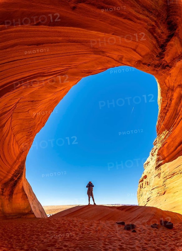 Person standing as a silhouette within the deep orange red sandstone walls of biidi the arch cave, looking out towards the bright blue desert sky and vast landscape of the american southwest