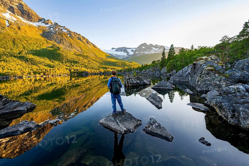 Young man standing on a rock in water, enjoying the landscape, picturesque mountain scenery in the Innerdalen high valley, mountain peaks reflected in Lake Innerdalsvatna, in the evening light at sunset, Trollheimen Mountain Area, Sunndal, Møre og Romsdal, Norway