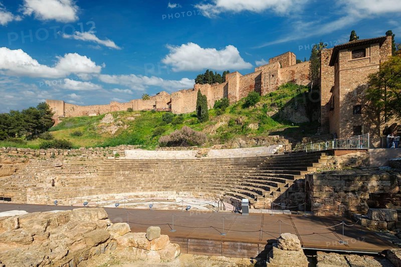 Scenic view of the Roman Theatre of Malaga and the Alcazaba walls across a green hillside, with ancient seating and stage ruins beneath sweeping white clouds, featuring roman ruins and spain