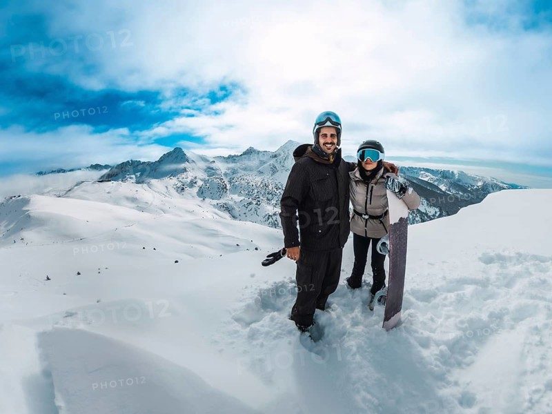 Smiling couple posing together with a snowboard on a snowy mountain summit, overlooking vast peaks and a vivid blue sky, embracing the thrill of winter sports and outdoor exploration
