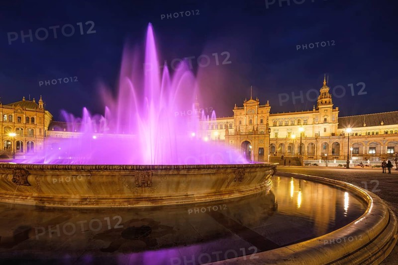 Purple illuminated fountain and curved basin stand before the glowing central pavilion of Plaza de Espana at night in Seville, Spain, featuring colored lighting