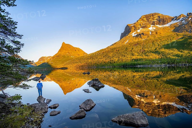 Young man in front of picturesque mountain landscape in the Innerdalen high valley with Innerdalstårnet summit, mountain peaks reflected in lake Innerdalsvatna, in the evening light at sunset, Trollheimen Mountain Area, Sunndal, Møre og Romsdal, Norway