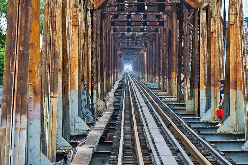 A Vietnamese State Railroad train crosses the Long Bien Railway Bridge across the Red River while a man crosses the tracks with a red bucket, Hanoi, Ha Noi province, Vietnam