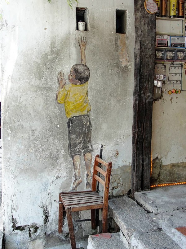 Street art titled Reaching Up by Ernest Zacharevic in Georgetown, Penang, Malaysia, travel photo, A mural of a child reaching high on a wall, with a chair nearby, set in an urban outdoor environment, George Town, Penang, Malaysia