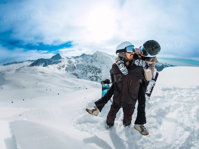 Snowboard couple sharing a playful piggyback on a snowy mountain peak, smiling and embracing winter adventure, scenic blue sky landscape and alpine joy