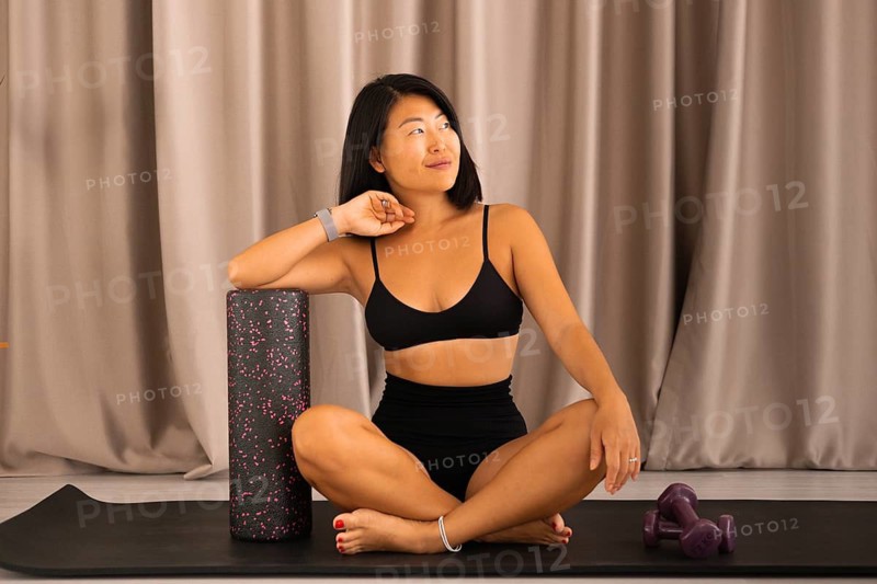 Woman sitting on a yoga mat with a foam roller and dumbbells in a calm indoor setting