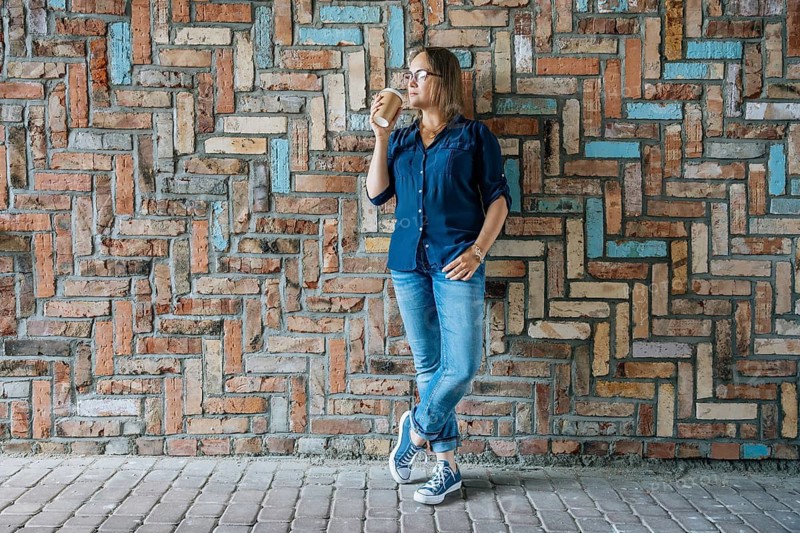 Woman drinking coffee while leaning against patterned brick wall, A woman in casual attire stands against a colorful brick wall, drinking coffee