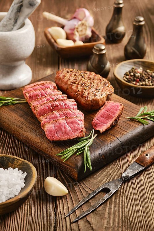 Beef steak sliced and arranged on wooden cutting board. Herbs, garlic and spices are placed around board