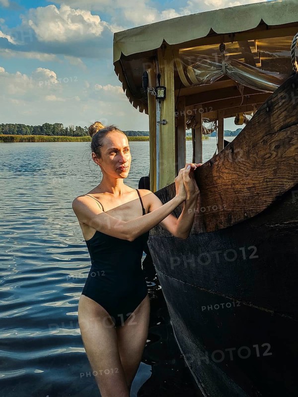 A young slim attractive woman in a one-piece swimsuit is relaxing by the lake and standing leaning against a wooden passenger vintage ship, A woman in a swimsuit stands beside a wooden boat on a sunny day by the lake