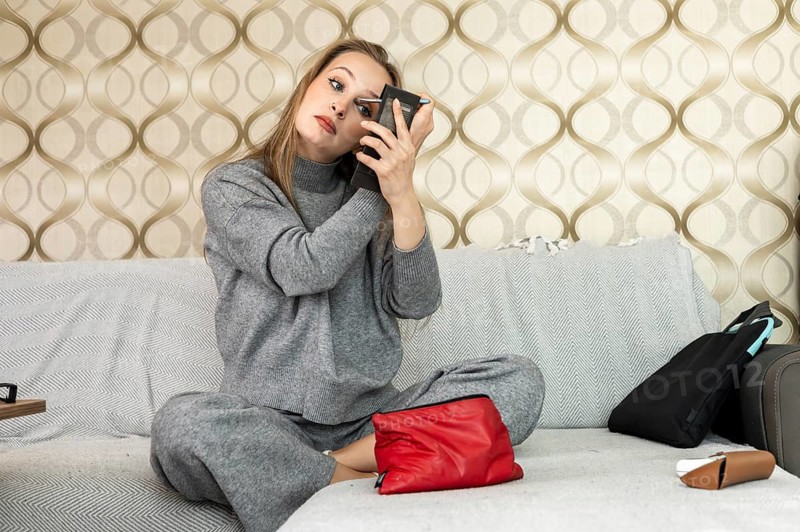 A young Caucasian woman with long brown hair applies makeup while sitting on a couch at home. She wears a gray outfit and looks focused in a handheld mirror. Home daily beauty routine