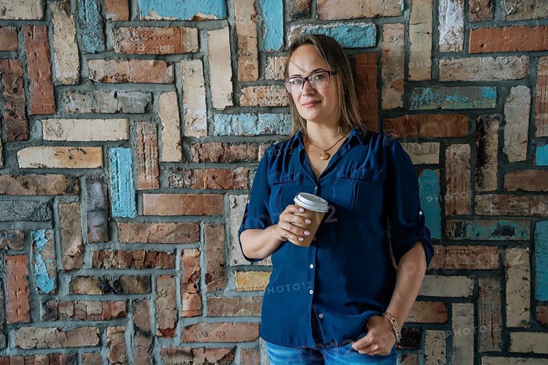 Woman holding a coffee cup, standing against a colorful brick wall. Casual attire, relaxed pose. Concept of leisure and comfort, A woman in a blue shirt stands with a coffee cup against a brick wall, exuding a casual and relaxed urban vibe