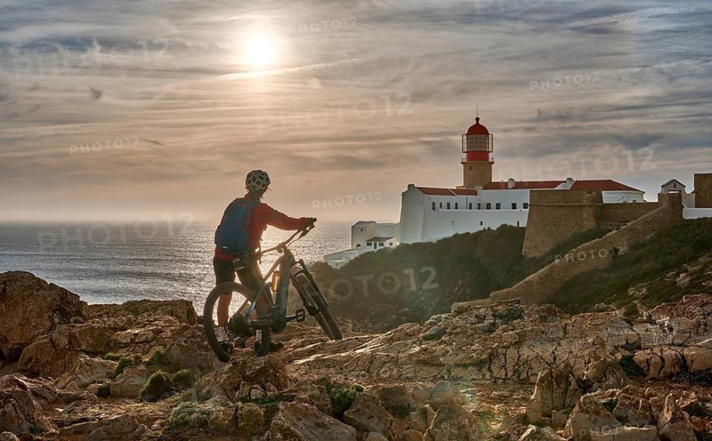Active senior woman cycling during moody golden hour near sunset at the the rock cliffs and lighthouse of Cabo Sao Vicente, the south-western spit of Europe at the atlantic coast, Sagres, Portugal