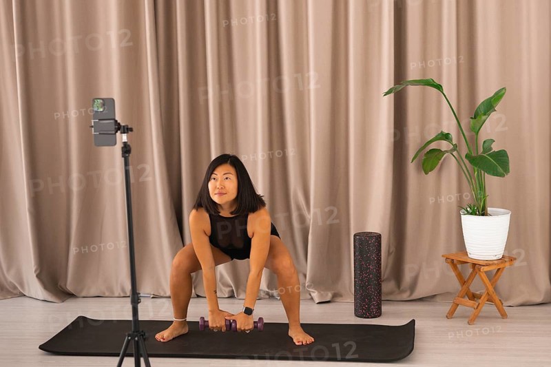 A woman squats with dumbbells, being recorded on a smartphone during an exercise session
