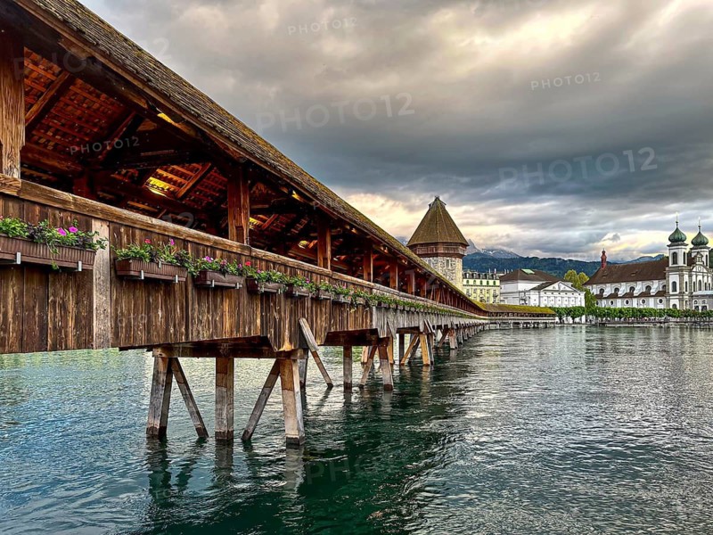 Wooden bridge leads across calm water surrounded by mountains and dramatic clouds, Chapel Bridge, Lucerne, Switzerland