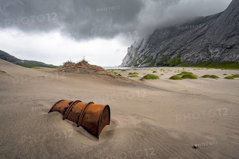 Rusted barrel between sand dunes on sandy beach Bunesstranden between rocky mountain peaks and dramatic clouds, Lofoten, Norway