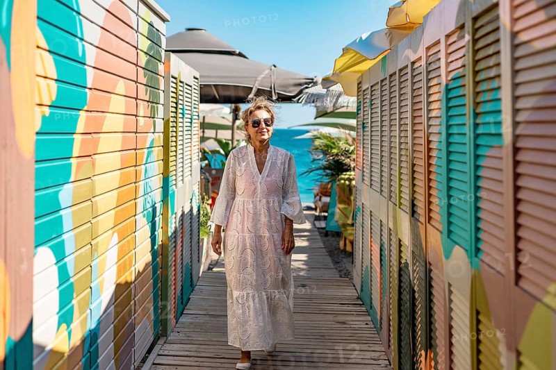 Mature Caucasian woman walks along a colorful boardwalk lined with beach huts and umbrellas. The scene is bright and sunny, evoking a relaxed summer atmosphere