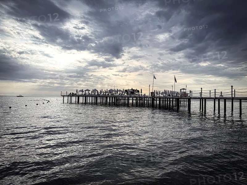 A long jetty with umbrellas stretches out into the calm sea under a dramatic sky, Side, Manavgat, Antalya Province, Turkey