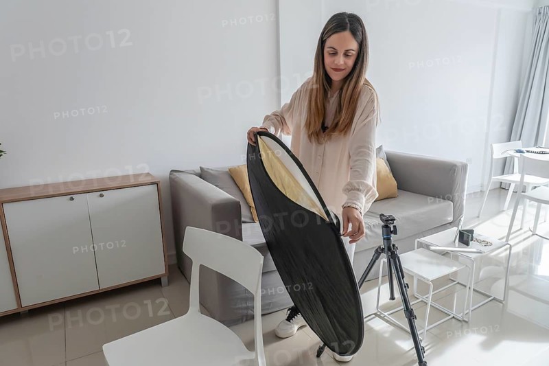 Woman standing and unfolding a collapsible gold and silver photography reflector beside a camera tripod in bright living room