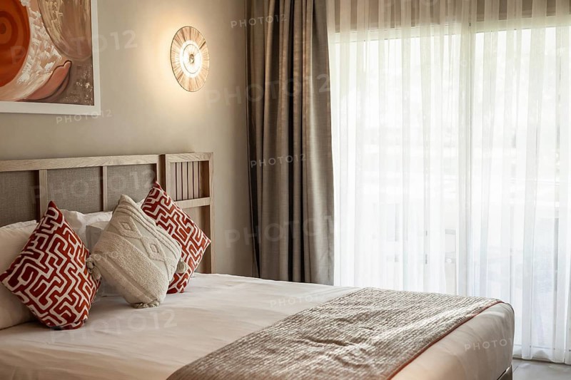 A modern hotel bedroom with a large bed, decorative pillows, and sheer curtains. The room features warm lighting and contemporary decor