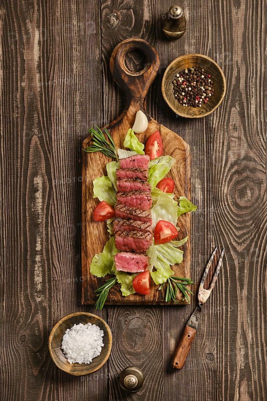 Beef steak cut into pieces placed on wooden cutting board. Plate includes lettuce, tomatoes, garlic and seasonings. Scene takes place in a kitchen with wooden elements and utensils