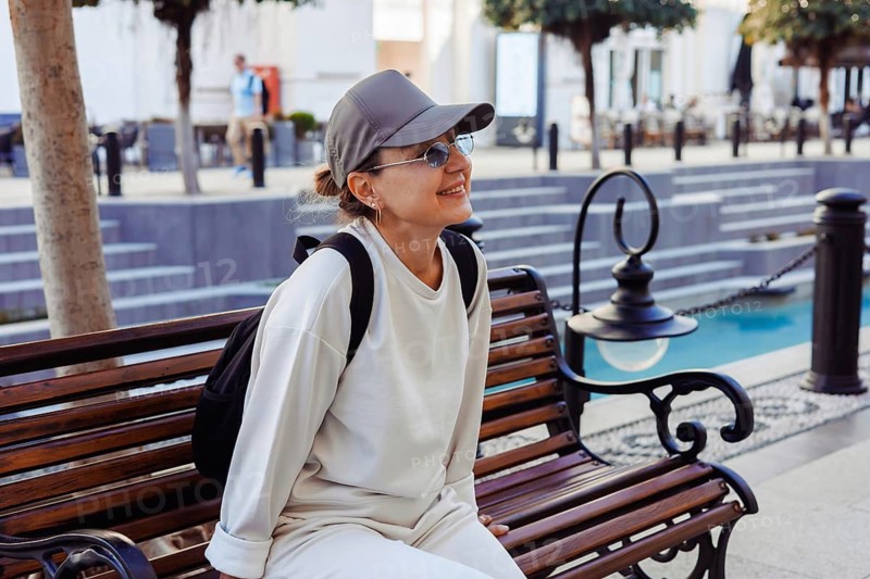 A cheerful middle-afed caucasian woman sits on a wooden bench, enjoying a sunny day in an outdoor area filled with trees and people. Shopping open air center, mall
