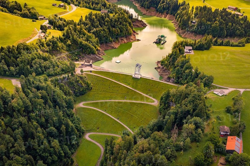 Aerial view of a winding road through green forest along a river with calm water, Krumbach, Vorarlberg, Austria